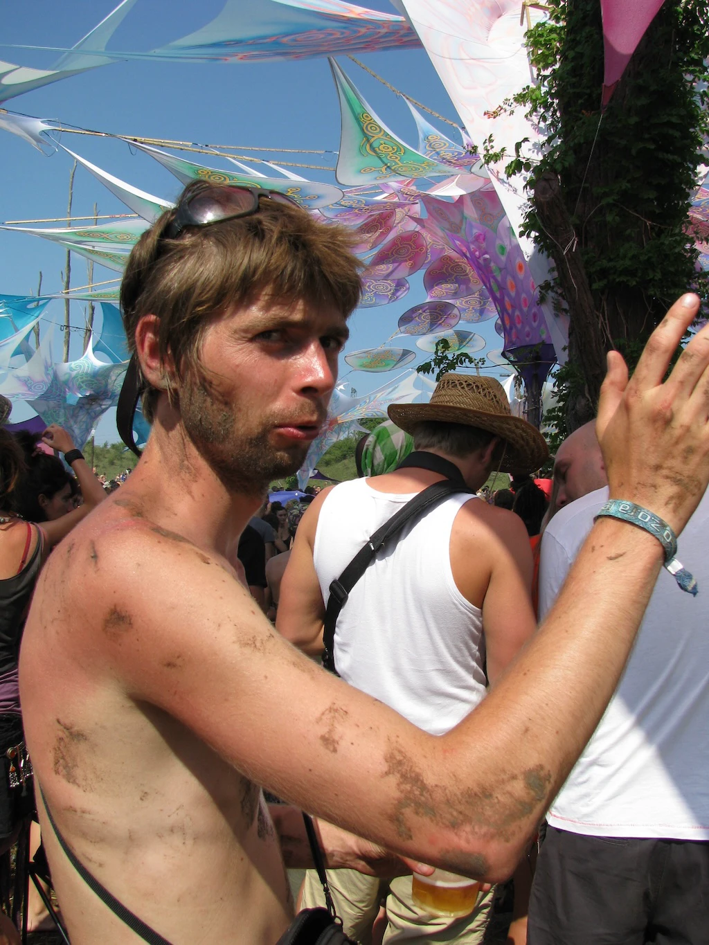 A shirtless man with dirt on his skin stands in a crowd at an outdoor festival, raising his hand. Colorful decorations hang above, and people are dancing around him.