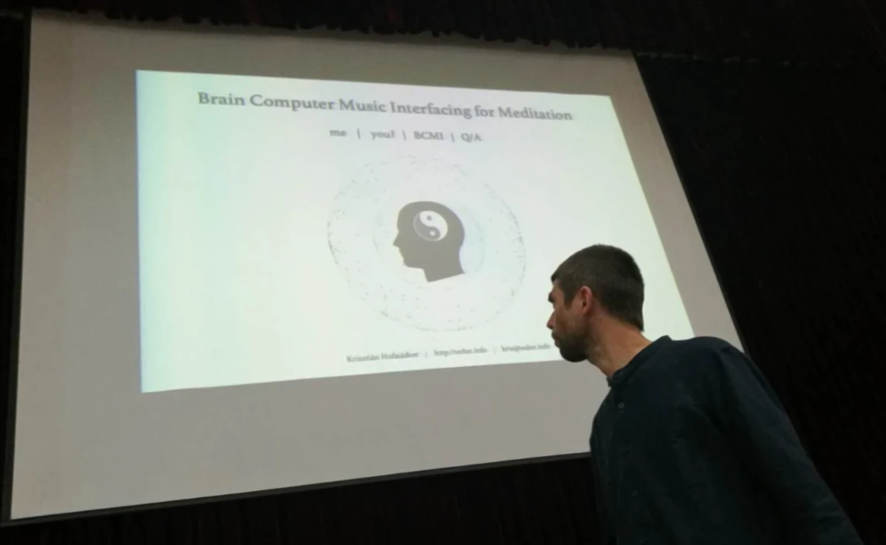 A speaker stands in front of a presentation slide titled "Brain Computer Music Interfacing for Meditation," featuring a yin-yang symbol and a head silhouette.